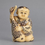 NETSUKE JAPONÊS EM MARFIM  Netsuke japonês esculpido em marfim, representando figura infantil sorridente trajando quimono decorado por motivos incisos e abraçando uma grande carpa. Medindo 5 x 3 cm, apresenta detalhes gravados com realces em pigmento escuro, além de passagens inferiores para cordão (himotoshi), próprias da função tradicional dos netsuke junto ao obi do quimono. A carpa é um motivo recorrente na tradição japonesa, associada à perseverança, força e prosperidade. Na base, conserva marca/inscrição em cartela vermelha.
