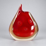 MURANO - Vaso escultórico em vidro artístico soprado. Medindo: 29 x 18 x 7 cm, peça com corpo assimétrico em formato de gota, núcleo vermelho intenso envolvido por espessa camada de vidro incolor e aplicações internas de bolhas ovais decorativas. A composição apresenta paredes espessas, base maciça e terminação superior alongada e curva, formando perfil orgânico característico do design italiano de vidro artístico do pós-guerra. A construção em camadas é compatível com a técnica sommerso, largamente empregada em Murano, na qual uma massa colorida é encapsulada por vidro de outra tonalidade ou por cristal incolor.