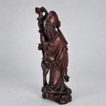 Escultura chinesa em madeira entalhada representando Shoulao (Shou Xing), divindade taoista associada à longevidade, medindo 25 x 9 x 8,5 cm, figurado como ancião barbado de longas vestes, segurando alto cajado de forma orgânica e acompanhado por frutos presos à indumentária. A iconografia de Shoulao é tradicionalmente associada ao cajado e ao pêssego da longevidade; exemplos comparáveis em madeira aparecem em leilões internacionais com essa mesma combinação de atributos. Christie's A peça apresenta entalhe em vulto pleno, barba longa, vestes de pregueado profundo e base integrada esculpida em forma de rochedos. O cajado sinuoso acompanha verticalmente a figura, reforçando a leitura tradicional do personagem.Shoulao integra o grupo dos chamados Três Deuses Estelares e simboliza longevidade; o pêssego constitui igualmente um dos seus atributos mais recorrentes na arte chinesa.
