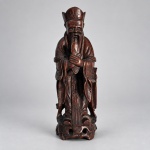 Escultura chinesa em madeira entalhada representando Lu Xing, divindade associada à prosperidade, posição social e êxito oficial, pertencente ao tradicional grupo Fu Lu Shou. Medindo: 21 x 7,5 x 6 cm. A figura aparece trajando longas vestes cerimoniais e barrete de oficial, segurando diante do corpo uma tábua cerimonial hu, atributo recorrente nas representações de Lu Xing e de altos oficiais chineses. Vinterior A peça apresenta execução em vulto pleno, com barba longa, vestes de pregueado marcado e base entalhada integrada à composição. A iconografia da tábua hu, originalmente vinculada à corte imperial e ao status oficial, reforça a identificação da figura no contexto das divindades auspiciosas chinesas.