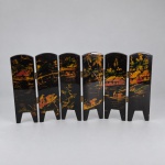 BIOMBO DE MESA ORIENTAL  Pequeno biombo articulado composto por seis folhas em madeira laqueada preta, decorado em ambas as faces. Medindo 19,5 x 36 cm aberto, apresenta em uma das faces paisagens asiáticas com casas, vegetação, embarcações e figuras em tons dourados, avermelhados e verdes. A outra face é ornamentada com aplicações de madrepérola, formando composições de aves, flores e folhagens sobre fundo preto. As seis folhas são unidas por pequenas dobradiças metálicas e terminam em pés recortados.