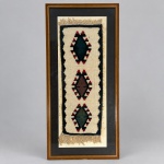 Pequeno kilim confeccionado manualmente em lã, com composição geométrica formada por três medalhões losangulares dispostos verticalmente sobre campo marfim, em tonalidades de verde, marrom, vermelho e preto. Medindo 83 x 38 cm com a moldura, apresenta cercadura escura, franjas nas extremidades e estrutura de trama plana característica dos kilims. A composição de medalhões geométricos alinhados encontra paralelo na tradição têxtil da Anatólia, Turquia, especialmente em exemplares de pequeno formato. Apresentado em moldura de madeira, sob vidro e com passe-partout escuro.