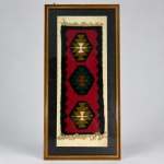 Pequeno kilim confeccionado manualmente em lã, com composição geométrica formada por três medalhões losangulares dispostos verticalmente sobre campo vermelho e cercadura preta. Medindo 83 x 38 cm com a moldura, apresenta motivos centrais em verde, ocre, branco e preto, além de franjas nas extremidades. A composição geométrica e a técnica de trama plana são características da tradição dos kilims da Anatólia. Apresentado sob vidro, com passe-partout escuro e moldura em madeira.