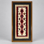KILIM ANATOLIANO  Pequeno kilim confeccionado manualmente em lã, com composição geométrica vertical sobre campo marfim, formada por sucessivos motivos florais e cruciformes estilizados em vermelho, azul, verde, marrom e preto, delimitados por cercadura vermelha e preta. Medindo 83 x 38 cm com a moldura, apresenta franjas nas extremidades e desenho simétrico de forte caráter geométrico, característico da tradição dos kilims da Anatólia. Apresentado sob vidro, com passe-partout escuro e moldura de madeira.