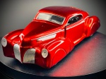 Miniatura Automóvel Clássico  Lincoln Zephyr Custom (Escala 1:43), Carroçaria em acabamento vermelho metalizado brilhante, portas e capô dianteiro/traseiro móveis (abríveis), interior detalhado com forras claras, motor visível sob o capô, pneu sobressalente alojado na bagageira, e chassis metálico inferior gravado com as especificações oficiais do fabricante e o selo de licenciamento. Marcas do tempo e de uso.
