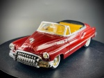 Miniatura Buick 1950 Cabriolet (Solido), com carroçaria em tom vermelho escuro (bordeaux), grelha frontal cromada de grande destaque, para-choques robustos com matrículas decorativas da Califórnia ("575 KIE"), interior detalhado em tons de amarelo/creme e pneus de faixa branca clássicos. O chassi inferior em metal exibe a gravação oficial do fabricante, referência de modelo (02-87), escala e origem. Marcas do tempo e de uso.