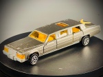 Miniatura Limousine (Cadillac)  Majorette, carroçaria em tom prateado/acinzentado com tejadilho em tom mais escuro, vidros e teto de abrir em acrílico âmbar, interior detalhado em amarelo, e um elemento decorativo em forma de boomerang fixado na tampa da mala traseira. O modelo dispõe de portas laterais operacionais. A base inferior exibe em relevo a marca Majorette, a escala 1/58, a designação Limousine e o local de fabrico (Made in France). Med. 02x13x03 cm. Marcas do tempo e de uso.