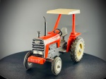 Miniatura de Trator Agrícola Massey Ferguson 275, rator agrícola clássico em tons de vermelho e cinzento, equipado com capota de proteção superior de cor clara com inscrições da marca, posto de condução aberto com assento e volante pretos, tubo de escape vertical e grelha frontal detalhada com emblemas alusivos ao modelo. "MASSEY FERGUSON 275" nas laterais do capô e na cobertura superior. Med. 06x08x05 cm.