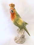 Estatueta de pássaro com coroa, repousando sobre tronco, em porcelana. Med. 21 cm.