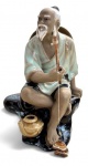 Escultura oriental em porcelana policromada representando ancião pescador sentado sobre rochedo, com vara de pesca, chapéu às costas e detalhes em relevo. Rica em acabamento e de caráter decorativo. Mede 14cm de altura.