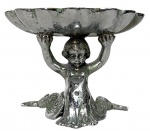 Saboneteira em metal prateado, em formato concha, sustentada por figura de putti (anjo) em relevo, com base decorativa. Peça de caráter ornamental, apresentando pátina e sinais do tempo. Mede 14cm x 10cm.