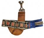 Adaga árabe antiga, possivelmente omanita ou iemenita, com lâmina curva em aço e empunhadura em madeira com aplicações decorativas. Acompanha bainha em couro com rica ornamentação têxtil bordada em padrões geométricos policromados. Peça de caráter etnográfico e decorativo, representativa da tradição armamentista do Oriente Médio. Mede 42 x 34 cm.