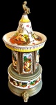 CAPODIMONTE. Antiga caixa de música em porcelana policromada, com rica decoração de cenas galantes e figuras em relevo, ornatos florais e detalhes dourados. Corpo guarnecido por elementos em metal patinado, apoiado sobre pés ornamentados. Parte superior arrematada por pequena figura de putto em metal, formando o puxador. Mecanismo musical funcionando. Apresenta discretíssimo bicado, conforme fotografia. Medidas: 35 x 17 cm de diâmetro.