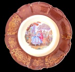 BAVARIA. Antigo prato decorativo em porcelana alemã, com aba recortada, ricamente ornamentada com arabescos e motivos florais em ouro. Centro com reserva policromada representando cena galante em jardim, com casal em trajes de época. Manufatura marcada no verso. Medidas: 27,5 cm de diâmetro.