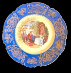 BAVARIA. Antigo prato decorativo em porcelana alemã, com aba recortada na tonalidade azul, ricamente ornamentada com arabescos e motivos florais em ouro. Centro com reserva policromada representando cena galante em jardim, com casal em trajes de época. Manufatura marcada no verso. Medidas: 28 cm de diâmetro.