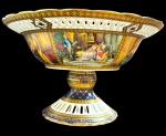 Imponente fruteira de centro em porcelana policromada, com rica ornamentação, sobre pedestal. Apresenta reservas decoradas com cenas galantes em interiores, intercaladas por frisos geométricos em azul-cobalto e ouro. Centro com composição floral policromada e ampla aba recortada e vazada. Medidas: 18 x 30,5 cm de diâmetro.