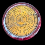 Calendário perpétuo de mesa, com mostrador circular em metal dourado e sistema giratório para consulta de datas, meses e anos, abrangendo o período de 1995 a 2034. Medidas: 2 x 7,7 cm.