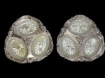 Wolff. Par de antigas petisqueiras em metal espessurado a prata, cada uma composta por três recipientes ovais em vidro prensado e lapidado, encaixados em estrutura de contorno recortado, ricamente ornamentada em relevo com volutas, concheados e motivos florais. Marca Wolff na base. Medidas: 23 x 24 x 2,5 cm cada.