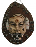 Imponente painel decorativo em madeira maciça, confeccionado a partir de seção transversal de tronco, preservando os veios e anéis naturais da madeira. Aplicado ao centro, máscara em relevo de inspiração veneziana, ornamentada com símbolos dos naipes de baralho, cartas, dados, peças de jogos e dominós distribuídos ao redor, formando composição temática ligada aos jogos de salão. Possui corrente para suspensão. Medidas: 55 x 42 x 8,5 cm.