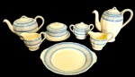 GRINDLEY, Cream Petal, England. Antigo jogo para chá e café em faiança, com decoração em faixas azul-celeste e frisos ornamentais em dourado sobre fundo marfim, composto por 7 peças: prato de bolo, dois bules, duas leiteiras, açucareiro e manteigueira.Medidas: prato de bolo: 27,3 x 24,5 x 3 cm; bule: 14,3 x 24,5 x 14,5 cm; leiteiras: 13,5 x 9 x 10 cm; manteigueira: 18 x 11 cm; açucareiro: 16 x 11 x 10 cm; bule maior: 23 x 13 x 20 cm. Dois bules apresentam pequenos bicados nos bicos.