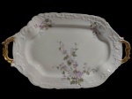 Antiga travessa/bandeja em porcelana tcheca, de formato retangular com cantos arredondados, bordas ricamente moldadas em relevo com motivos rocaille e duas alças laterais vazadas. Decoração floral policromada em delicados ramos de flores em tons de lilás e folhagens, com detalhes dourados nas alças. Marcada na base.  Medidas: 39 x 24,5 x 4 cm.