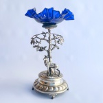 Belo centro de mesa em metal alemão prateado. Base adornada com cervo embaixo de árvore sobre quatro pés e encimado por prato na tonalidade azul cobalto em babados.Falta um pedaço do chifre. Med 30 cm. 