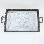 Grande tabuleiro para banquete em metal espessurado à prata com galeria vazada e alças laterais com decoração floral, fundo lavrado com volutas e elevada por 4 pés. Med 73 x 41 x 7 cm. 