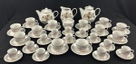 SCHMIDT. Aparelho de chá e café para 12 pessoas, confeccionado em porcelana branca, com delicada decoração floral de rosas, composto por 54 peças: 12 xícaras de café com respectivos pires, 12 xícaras de chá com respectivos pires, bule de café, bule de chá, leiteira, manteigueira, açucareiro e prato para bolo. Sem uso, acondicionado em caixa original.