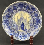 Prato decorativo em faiança, com estampa monocromática em azul, representando figura religiosa diante de catedral em estilo Neogótico, circundada por elaborada ornamentação floral e arabescos. Medida: 21 cm de diâmetro.