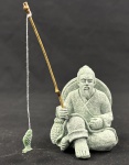Pequena escultura oriental em material de aspecto pétreo, representando pescador sentado, acompanhado de cesto, vara de pesca em bambu e peixe pendente. Corpo com acabamento texturizado em tonalidade verde-acinzentada. Medidas da escultura: 8 x 6 cm.