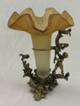 Vaso decorativo estilo épergne, apresentando tulipa em vidro acetinado fosco na tonalidade âmbar com borda superior ondulada e frisada, sustentado por estrutura escultórica em metal fundido, ornado com motivos florais e uma figura de querubim (putto). Medindo: 24 cm.
