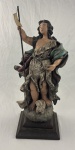 Escultura sacra representando São João Batista, em pesado barro cozido de feição expressiva e composição dinâmica, inspirada na estética barroca, com rico detalhamento nas vestes, movimento acentuado e policromia em tons terrosos e esverdeados, portando estandarte, apoiado sobre base trabalhada com elementos simbólicos. Assinada Tiana e datada de1987. Medindo: 62 x 24 x 21 cm