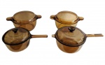 VISION. Conjunto de quatro panelas em vidro temperado âmbar translúcida, sendo duas com cabos alongados e duas com alças laterais, todas acompanhadas das respectivas tampas. Uma das tampas apresenta pequeno bicado na borda. Medidas: 9 x 31 x 18 cm; 17 x 37 x 21,5 cm; 14 x 30,5 x 21,5 cm.