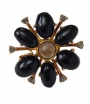Anel e imponente em metal dourado, de formato floral, ornamentado por sete pedras negras de lapidação cabochão, pedra central  facetada e pequenas pedras incolores cravejadas nas extremidades. Aro;178