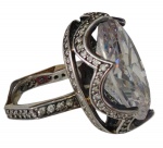 Grande e imponente anel em prata de lei 925, ornamentado por volumosa pedra incolor de lapidação oval facetada, sustentada por garras e circundada por aro vazado de desenho ondulado, cravejado por pequenas pedras incolores, que se prolongam pelas laterais. Aro: 17. Peso: 28,9 g.