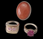 Três anéis em prata, sendo um ornamentado por grande pedra oval em tom alaranjado, de lapidação cabochão e efeito cintilante; um de aro largo, com duas pedras rosadas de lapidação oval facetada; e um com pedra central em tom rosa intenso, ladeada por pequenas pedras incolores cravejadas. Aros: 22, 19 e 19. Peso total: 26,9 g.