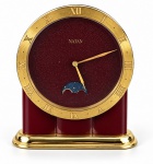 RELÓGIO, um (1) relógio de mesa NATAN, suíço, com caixa esmaltada em tom vinho e acabamento dourado, mostrador circular vinho com algarismos romanos aplicados em dourado, ponteiros dourados e decoração inferior em forma de lua crescente azul com estrelas. Base dourada de formato retangular arredondado. Peça em excelente estado de conservação, acondicionada em estojo original, não testado e sem garantia de funcionamento, medindo 13x11x10,5cm.
