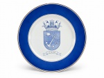 PORCELANA P II, um (1) prato decorativo confeccionado em porcelana na tonalidade branca, decorado com ampla faixa em azul cobalto, filetes dourados e ao centro brasão de inspiração naval/militar, com âncora, canhão e engrenagem, acompanhado da sigla CAM, apresentando falhas na douração, medindo 26cm de diâmetro.