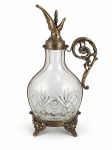 CRISTAL, uma (1) licoreira confeccionada em cristal translucido, com corpo bojudo com lapidação geométrica e de palmas em baixo relevo, guarnições em bronze trabalhado em relevo, alça lateral ornamentada, base com pés e tampa decorativa com bico vertedor. Medida :  32 cm , possui pequeno trincado no gargalo .