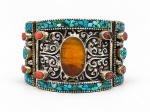 BIJUTERIA, um (1) bracelete rígido em alpaca, estilo tibetano, ricamente trabalhado com motivos vegetalistas em relevo, aplicação central oval em possivelmente âmbar e ornamentação com pedrarias em tons de turquesa e coral, incluindo trabalho em cascalhos de turquesa, sinais de uso, vendido conforme a foto, medindo 7,5cm de diâmetro.