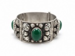 BIJUTERIA, um (1) bracelete articulado em metal prateado, de inspiração oriental, composto por placas trabalhadas em relevo com motivos florais e aplicações de cabochões em possivelmente ágata verde, medindo 6,5cm de diâmetro.