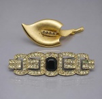 BIJUTERIAS, dois (2) broches, sendo: A) 1 broche em metal dourado, em formato de folha estilizada, com aplicação central de zircônias, sinais de uso, medindo 6,5x3cm e B) 1 broche em metal prateado da marca CAROL, desenho geométrico estilo Art Déco, cravejado com zircônias e pedra central preta facetada, sinais de uso, medindo 8x2cm.