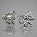 BIJUTERIAS, dois (2) broches confeccionados em metal espessurado a prata, sendo: A) 1 broche de desenho floral e texturizado em vazados, incrustado por zirconias e pérola sintética central, medindo 4,5x5cm e B) 1 broche em formato de flor estilizada, com pétala vazada e aplicação de zircônias, em vazados, medindo 6,5x6cm.