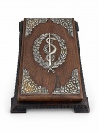 PRATA DE LEI, um (1) porta bloco de notas em jacarandá com adornos florais nos cantos e ao centro o bastão de Asclépio (símbolo da medicina) em PRATA DE LEI, com pequena falha no adorno inferior direito, medindo 4,5x25x17cm.