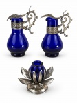 METAL PRATEADO e VIDRO, três (3) peças confeccionadas em vidro na tonalidade azul cobalto, sendo: A) 2 galhetas com cinzelado de cachos de uva e folhas de parreira em relevo, guarnição em elementos florais em vazados e alça em arabesco vazado, falhas na prateação, medindo 12cm e B) 1 castiçal no feitio de flor confeccionado em metal espessurado a prata com falhas na prateação e mossa na base, recipiente em vidro na tonalidade azul cobalto, medindo 7x9,5cm.