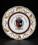 PORCELANA REAL, um (1) prato decorativo confeccionado em porcelana na tonalidade branca,  centro decorado por brasão e a insígnia do Centro de Instrução do Corpo de Fuzileiros Navais da Marinha do Brasil em policromia com elementos vegetalistas em ouro e espessa faixa marrom, medindo 25cm de diâmetro, leves falhas.
