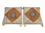 CAMA, MESA & BANHO, duas (2) capas de almofada em tecido tipo brocado/sedoso, com padronagem floral e aplicação central em losango na cor laranja, com detalhe circular ao centro, acabamento com pingentes/franjas nos cantos, medindo 40x40cm.