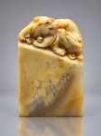 ARTE ORIENTAL, um (1) selo de carimbo chinês esculpido em pedra, possivelmente pedra Shoushan, em tonalidades amarelas, creme e acastanhadas, com corpo retangular e topo ricamente entalhado com criatura mítica chinesa, semelhante a leão-guardião (Fo/Foo), acompanhada de figura menor. Peça de tradição oriental, com variações naturais de coloração e veios da pedra, pequeno bicado na base, medindo 6x4x2,5cm.