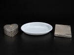 METAL PRATEADO e PORCELANA, lote com três (3) peças, sendo: A) 1 caixa porta joias confeccionada em metal prateado no formato de coração, decorada por elementos vegetalistas e coração cercado por perolados, interior em tecido vermelho, medindo 3,5x7x6,5cm, B) 1 travessa oval da marca SCHMIDT, confeccionada em porcelana na tonalidade branca, medindo 17x11,5cm e C) 1 cigarreira confeccionada em metal prateado, cinzelado de ondulações e placa retangular em reserva, sinais de oxidação, medindo 10x9cm.