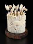 DIVERSOS, um (1) conjunto porta-palitos decorativo, composto por recipiente cilíndrico em material sintético de tonalidade marfim, entalhado em relevo com figura de animal e motivos vegetais, acompanhado de onze palitos com terminações esculpidas em formas de folhas, flores e aves. Base circular em madeira. Peças no estado, apresentando fios de cabelo, medindo 9,5x10cm.